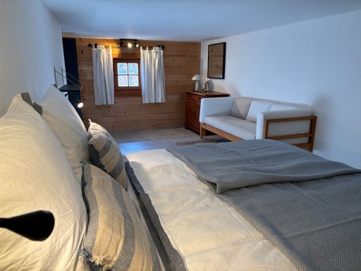 "Scheunenschlafzimmer" Helles Schlafzimmer mit Doppelbett, Sofaecke und Fenster mit Vorhängen.