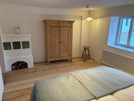 "Stubenschlafzimmer" Helles Schlafzimmer mit Holzfußboden, Kachelofen, Kleiderschrank und Fenster.